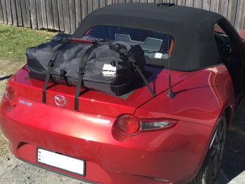red mazda miata mx5 nd with a boot-bag original luggage rack fitted to the boot/trunk outside next to a fence photographed from the rear